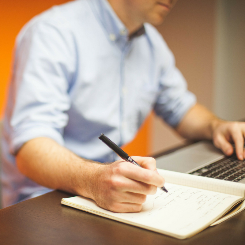 man sitting at laptop and writing in notebook
